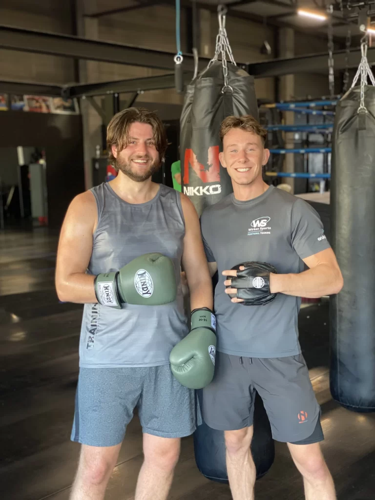 Twee mannen poseren in sportschool met bokshandschoenen.